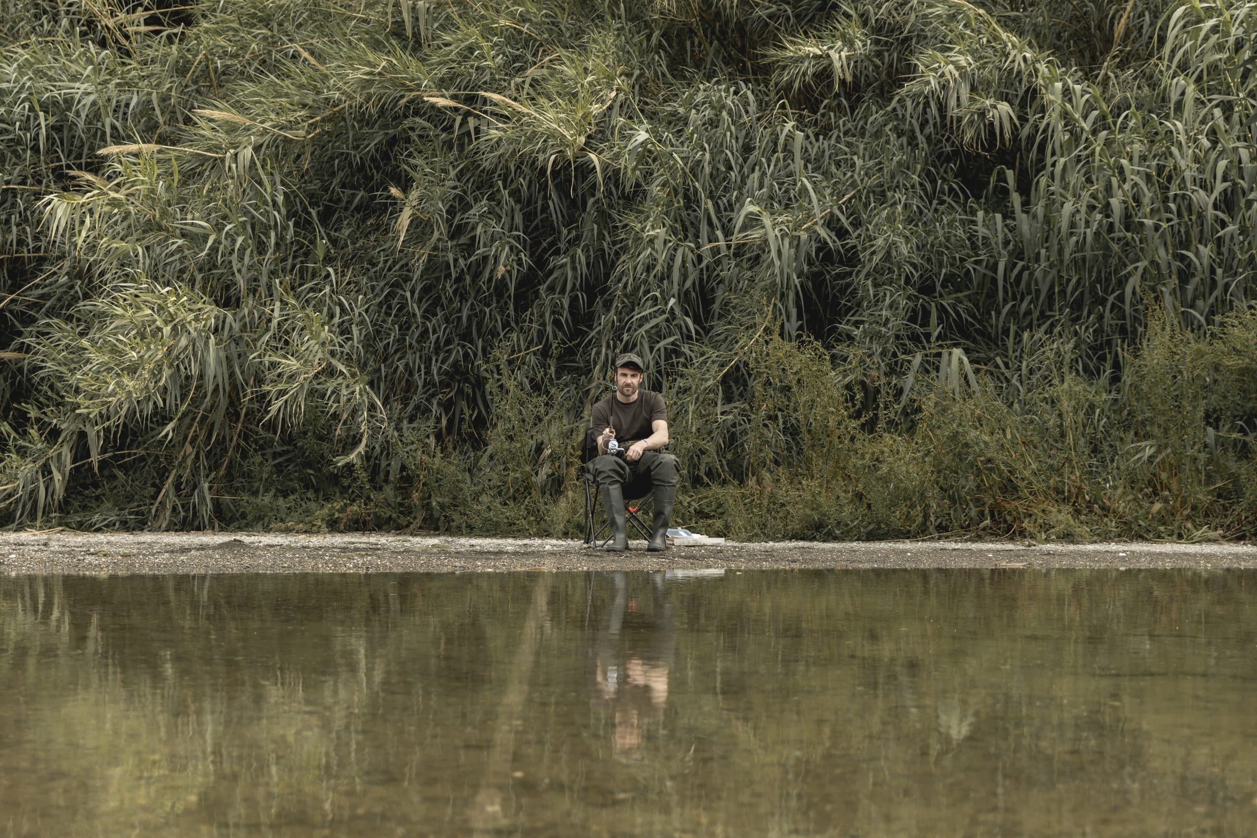 Angler seated on a low stool at a reed-lined riverbank, rod in hand near a calm stretch of water